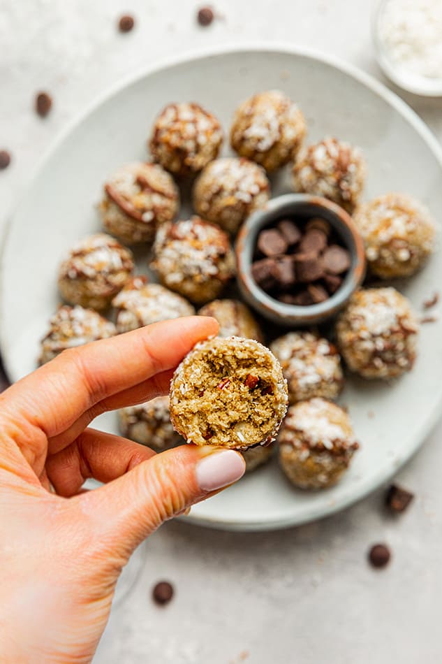 A hand holding a coconut protein ball in front of a plate of protein balls