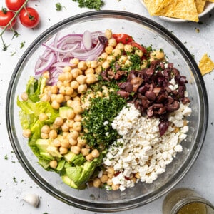 Overhead view of chopped cucumber, tomatoes, bell peppers, avocado, red onions, chickpeas, parsley, olives and feta side-by-side in a large clear mixing bowl to make a Mediterranean Chickpea Salad