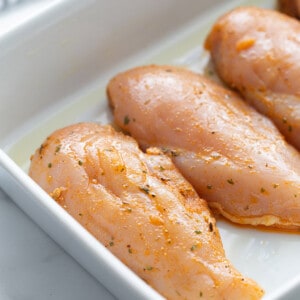 Side shot of four raw chicken breasts in a white rectangle baking dish