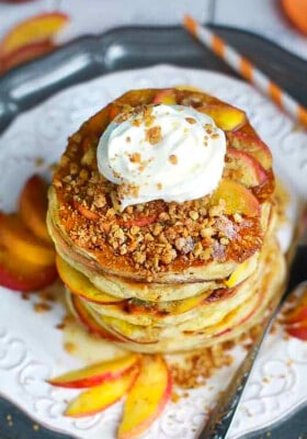 Overhead shot of a stack of six fluffy peach pancakes topped with coconut whipped cream and sliced peaches on a white plate