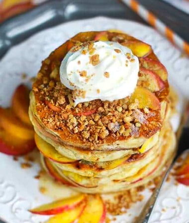 Overhead shot of a stack of six fluffy peach pancakes topped with coconut whipped cream and sliced peaches on a white plate