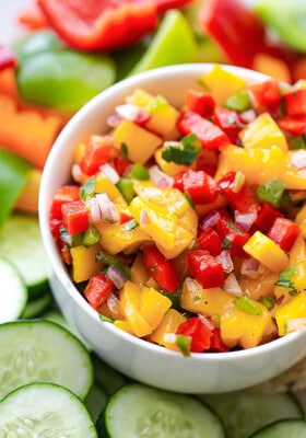 Side shot of a serving of fresh peach salsa in a white bowl with sliced cucumbers
