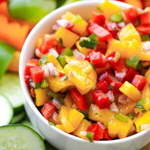 Side shot of a serving of fresh peach salsa in a white bowl with sliced cucumbers