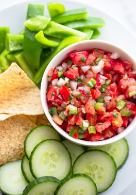Top close-up shot of a serving of fresh tomato salsa in a white bowl with sliced cucumbers, bell peppers and tortilla chips