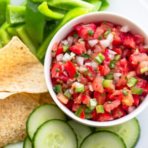 Top close-up shot of a serving of fresh tomato salsa in a white bowl with sliced cucumbers, bell peppers and tortilla chips