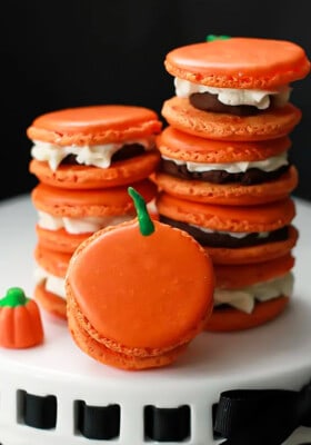 Eight pumpkin macarons stacked in two piles with one in front on a white cake stand.