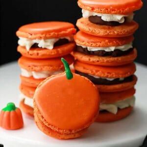Eight pumpkin macarons stacked in two piles with one in front on a white cake stand.