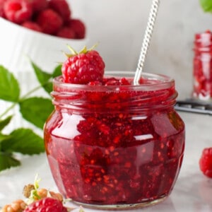 Side shot of a jar of raspberry chia jam with a spoon in the jar and raspberries on the side