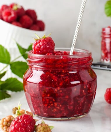 Side shot of a jar of raspberry chia jam with a spoon in the jar and raspberries on the side
