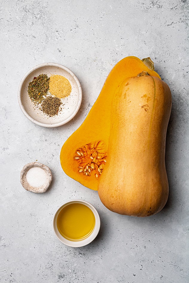 A halved butternut squash on a kitchen countertop beside avocado oil, sea salt and the remaining ingredients