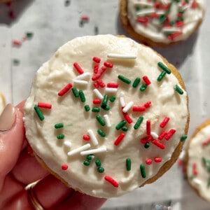 A hand holding one iced sugar cookie with green and red sprinkles over white parchment paper.