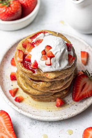 45 degree angle of a stack of gluten free strawberry pancakes topped with sliced and diced strawberries with whipped cream on a white plate