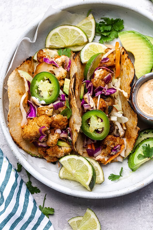 Overhead view of cauliflower tacos on a plate with limes, avocado, and jalapeno slices