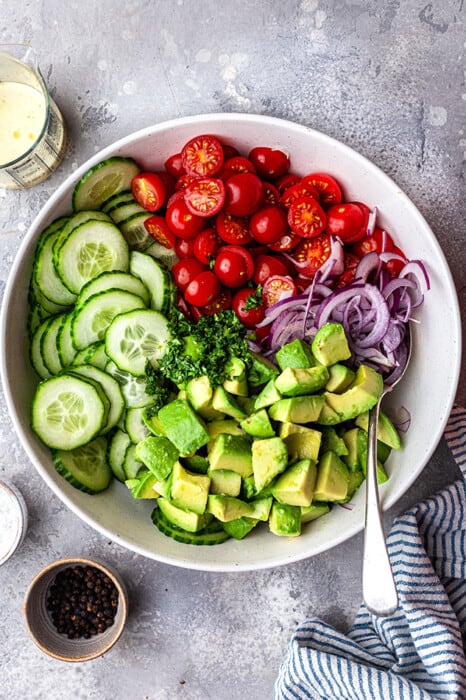 A bowl of cucumbers, cherry tomatoes, red onion and avocado in a white bowl