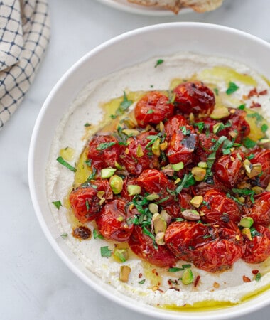 Overhead top shot of roasted cherry tomatoes piled on whipped feta in a white bowl with some crusty bread on the side