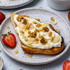 A piece of ricotta toast topped with sliced walnuts and almonds on a plate with a halved strawberry