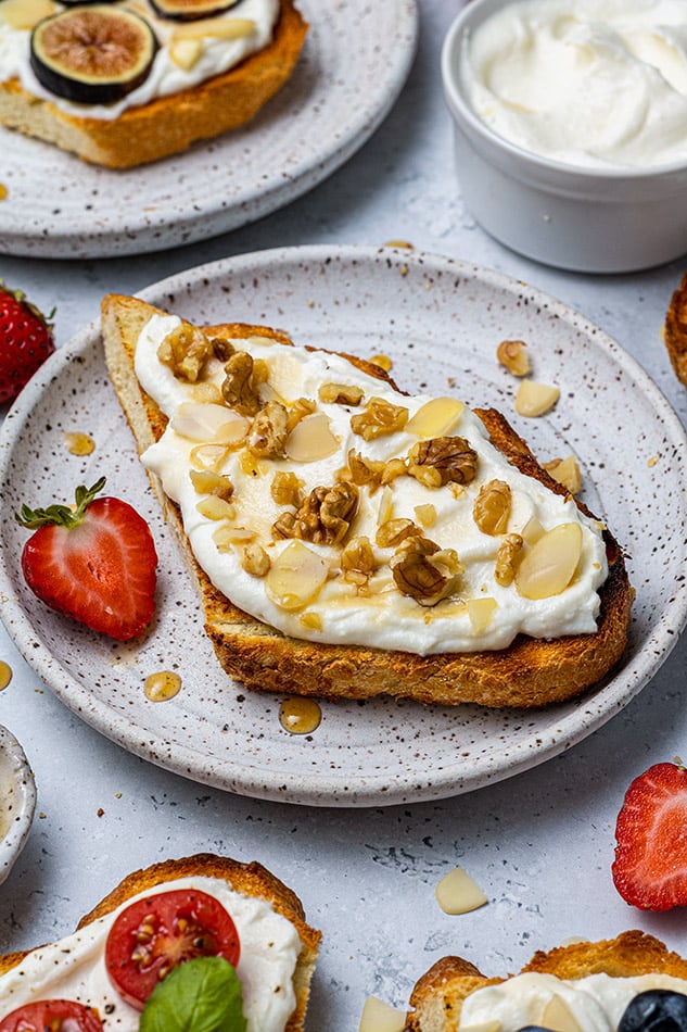 A piece of ricotta toast topped with sliced walnuts and almonds on a plate with a halved strawberry