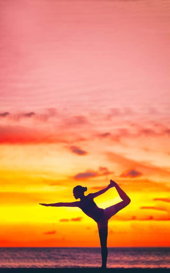 Yoga silhouette of woman stretching doing dancers pose in beautiful dusk colors at sunset on beach background. Copy space on pink sky clouds. Wellness meditation concept.