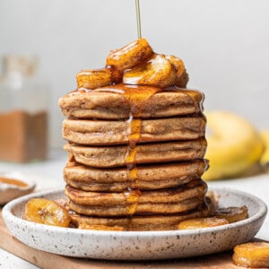 A tall stack of gluten-free banana pancakes with maple syrup being poured on top