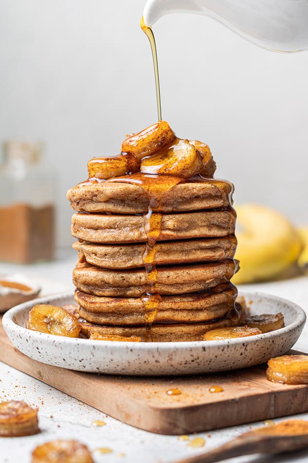 A tall stack of gluten-free banana pancakes with maple syrup being poured on top