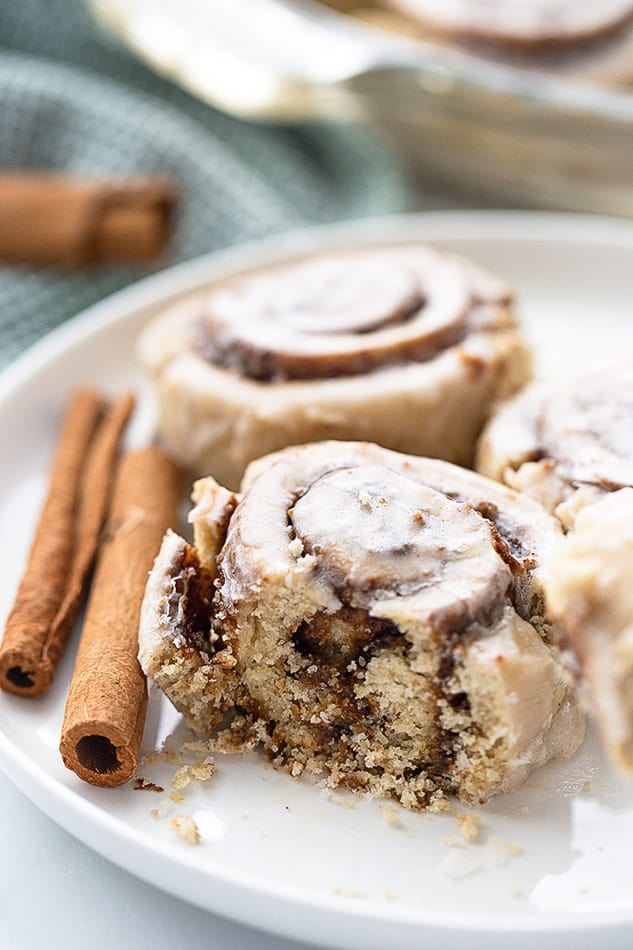 Close-up of a cinnamon roll on a plate with cinnamon sticks and two more cinnamon rolls in the background