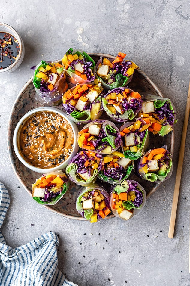 Top view of 14 fresh summer rolls in a large beige bowl with peanut dipping sauce and chopsticks