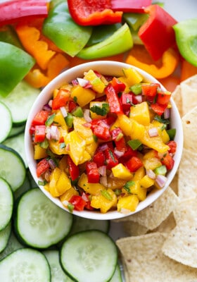 Top shot of a serving of fresh mango salsa in a white bowl with sliced cucumbers, bell peppers and tortilla chips