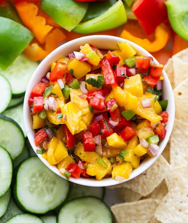 Top shot of a serving of fresh mango salsa in a white bowl with sliced cucumbers, bell peppers and tortilla chips