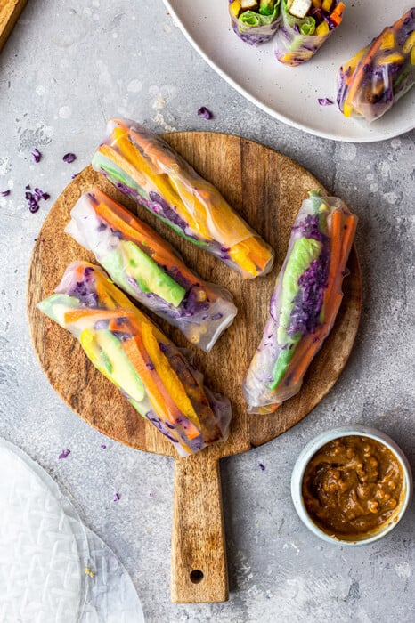 Four freshly wrapped summer rolls on a cutting board with a couple of halved rolls on a plate beside it