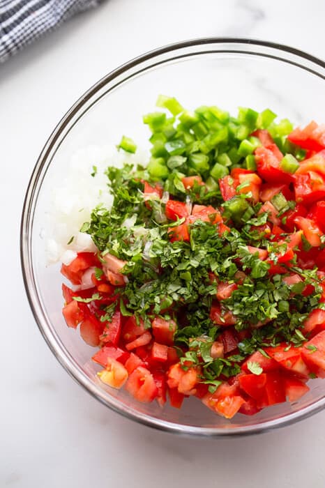 A clear mixing bowl filled with ingredients to make the tomato salsa