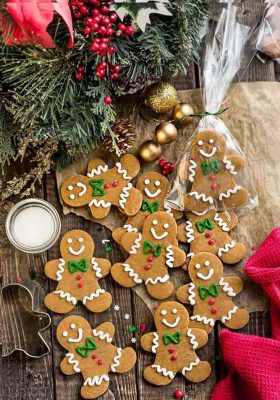 Top view of a batch of decorated gingerbread men cookies on a wooden board surrounded by a mistletoe and bells.