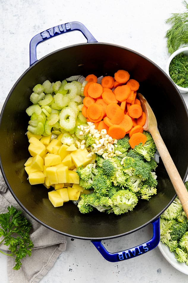 Chopped carrots, celery, diced potatoes, broccoli, garlic and onions in a blue dutch oven / pot with a wooden spoon