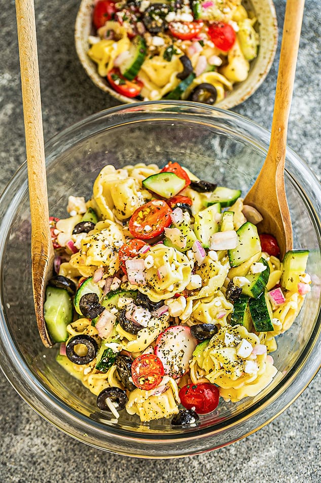 A clear mixing bowl with gluten free tortellini pasta salad with two wooden salad tossers