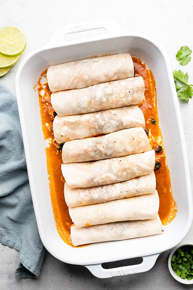 Eight wrapped enchiladas in a baking dish with a layer of enchilada sauce on the bottom