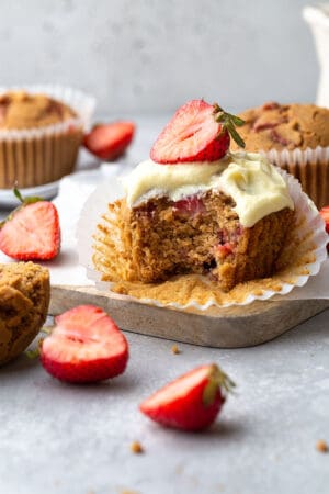 Side view of one gluten-free strawberry muffin topped with whipped coconut cream with a missing bite on a grey background