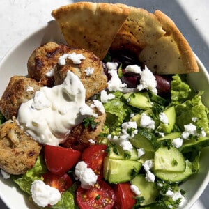A serving of chicken meatballs over a Greek salad with pita and feta in a white bowl