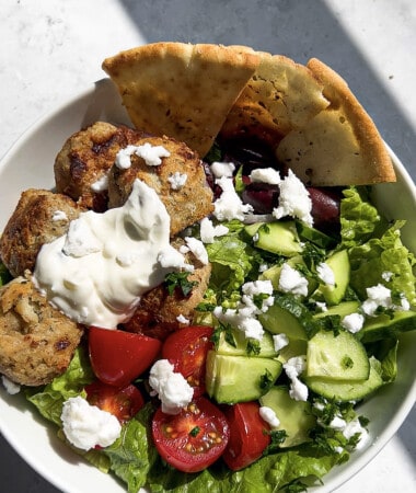 A serving of chicken meatballs over a Greek salad with pita and feta in a white bowl