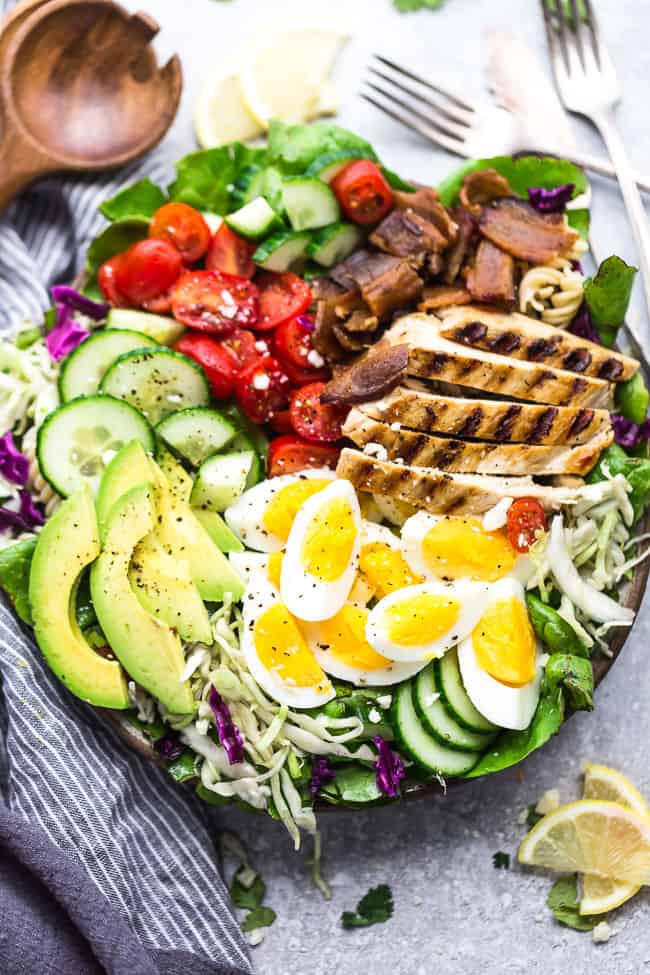 The top view of a Cobb salad on a gray surface beside lemon slices and a wooden spoon