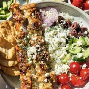 A serving of 2 greek chicken skewers over white rice with cherry tomatoes, cucumbers, kalamata olives, red onion, feta and pita in a white bowl