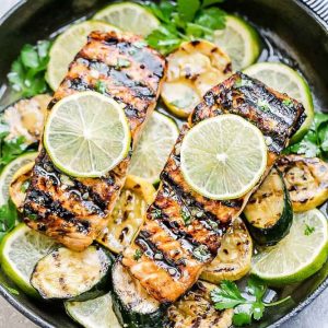 Top view of two Honey Lime Salmon fillets in a skillet with sliced zucchini and limes