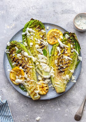 Overhead view of three grilled romaine lettuce halves on a grey plate topped with vegan Caesar dressing with lemon halves