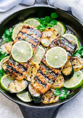 A Top View of Two Garlic Lime Salmon Fillets in a Skillet with Sliced Zucchini and Limes