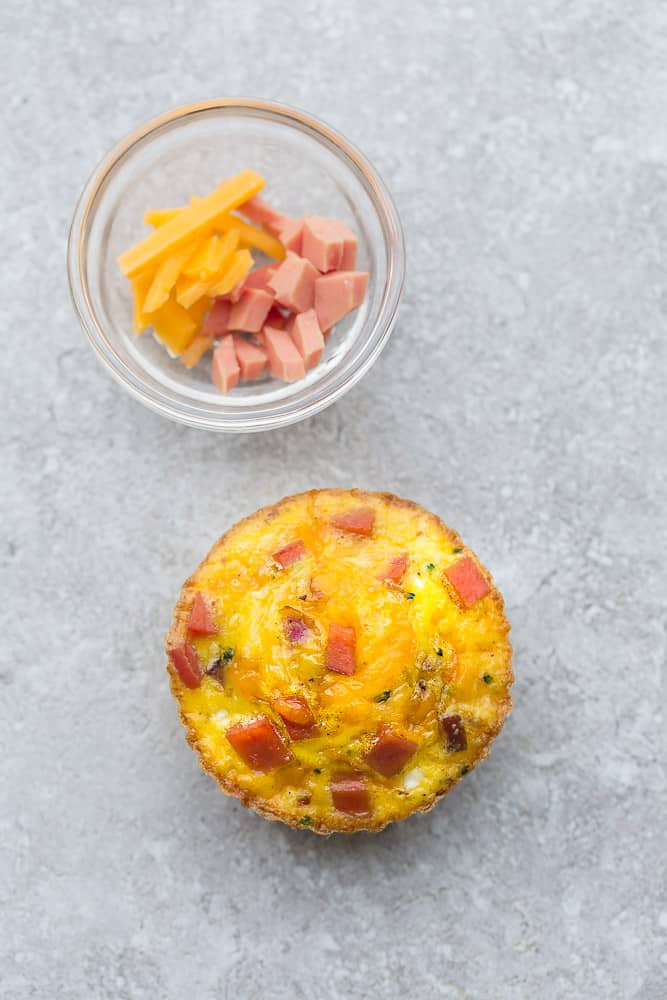 A Ham and Cheese Egg Muffin on a Gray Surface Beside a Small Dish of Shredded Cheddar Cheese and Ham