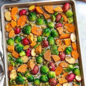 Flat lay of chopped roasted sweet potatoes, potatoes, Brussels sprouts, carrots and seasonings on a large baking sheet.