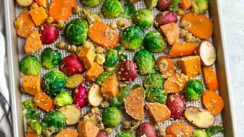 Flat lay of chopped roasted sweet potatoes, potatoes, Brussels sprouts, carrots and seasonings on a large baking sheet.