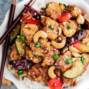 A bowl of kung pao chicken over rice with a pair of chopsticks balancing across the rim of the bowl