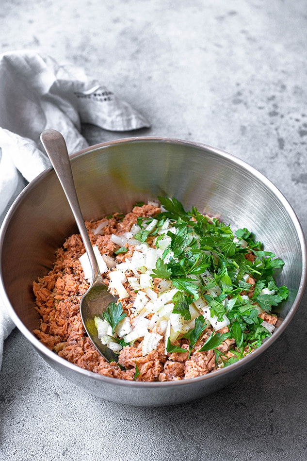 Flaked fish in a metal bowl with diced onion, chopped parsley and a mixing spoon