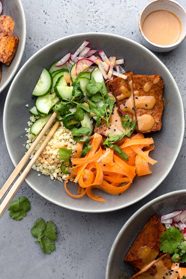 A serving of vegan bánh mi bowl topped with ribboned carrots, cauliflower rice, crispy tofu and fresh herbs in a grey bowl with chopsticks in the bowl