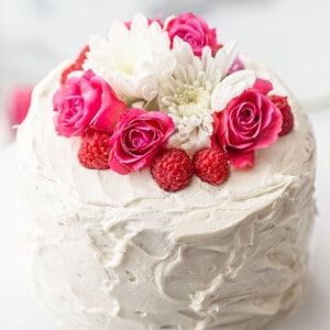 A whole Keto Vanilla Cake topped with fresh flowers and raspberries on a white cake stand
