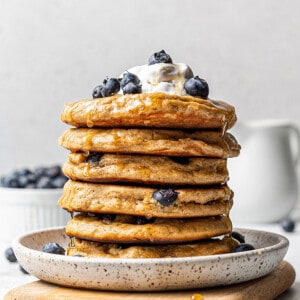Side shot of a stack of five healthy blueberry pancakes topped with blueberries with whipped cream on a white plate